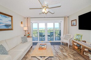 A living room with a sofa, a coffee table, a ceiling fan, a TV, and a balcony with outdoor furniture. - Fabulous views from this Vibrant Oceanfront Townhouse -Sea Place 15274! (St. Augustine)