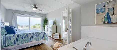 Een slaapkamer met een bed, een plafondventilator, een raam met gordijnen, een dressoir en een schilderij van een strand.