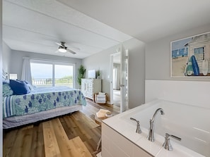 A bedroom with a bed, a ceiling fan, a window with curtains, a dresser, and a painting of a beach scene.