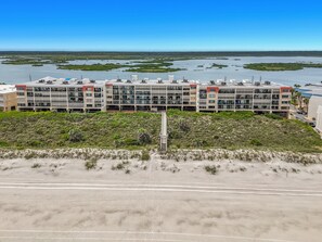 Ein mehrstöckiges Hotel mit flachem Dach, in Strandnähe, mit Sanddünen und einem Gewässer im Hintergrund.