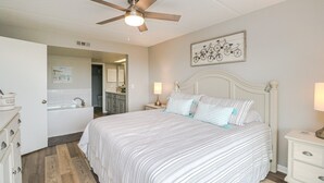 Une chambre à coucher comprenant un lit, des tables de chevet, un ventilateur de plafond, une salle de bain avec un lavabo et un miroir, et une œuvre d’art représentant un vélo, accrochée au mur.