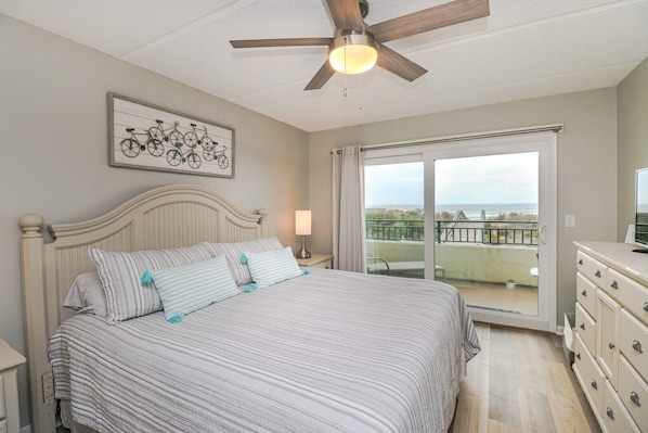 A bedroom with a bed, a ceiling fan, a nightstand, a lamp, a dresser, and a balcony with a view.