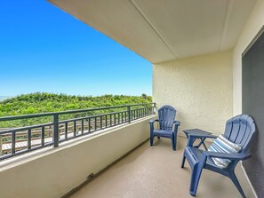 A balcony with two chairs and a small table, overlooking a lush green landscape and the sea. - Windjammer 109 - Comfortable Ocean View Condo, 2BR, Great Rates, Elevator & Covered Parking (St. Augustine)
