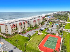 Un hotel frente a la playa con edificios de varios pisos, una cancha de tenis y un estacionamiento.