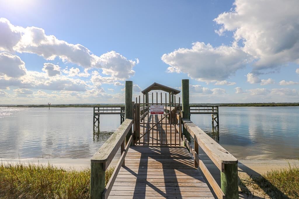 Eine Holzstege führt zu einem überdachten Bereich mit einem Schild, umgeben von ruhigem Wasser und blauem Himmel.