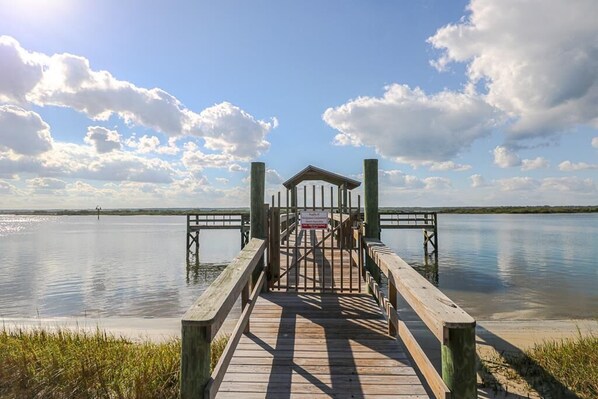 A wooden pier leading to a covered area with a sign, surrounded by calm water and a clear sky.