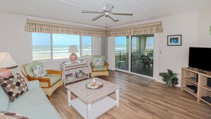 A living room with a ceiling fan, wicker furniture, a television, and a view of the beach. - Windjammer 212 - Direct Oceanfront, Intracoastal Waterway Views in Crescent Beach (St. Augustine)