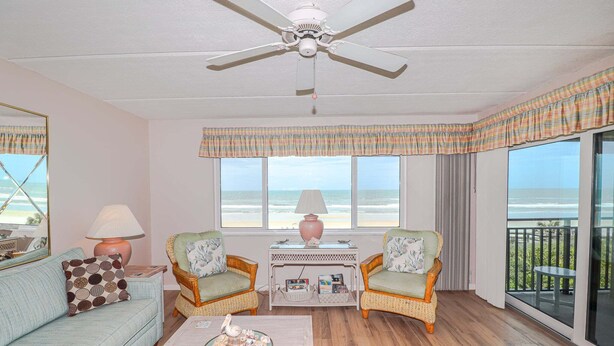 A room with a ceiling fan, two wicker chairs, a sofa, a table with a lamp, a window with a view of the beach, and a balcony with a table. - Windjammer 212 - Direct Oceanfront, Intracoastal Waterway Views in Crescent Beach (St. Augustine)