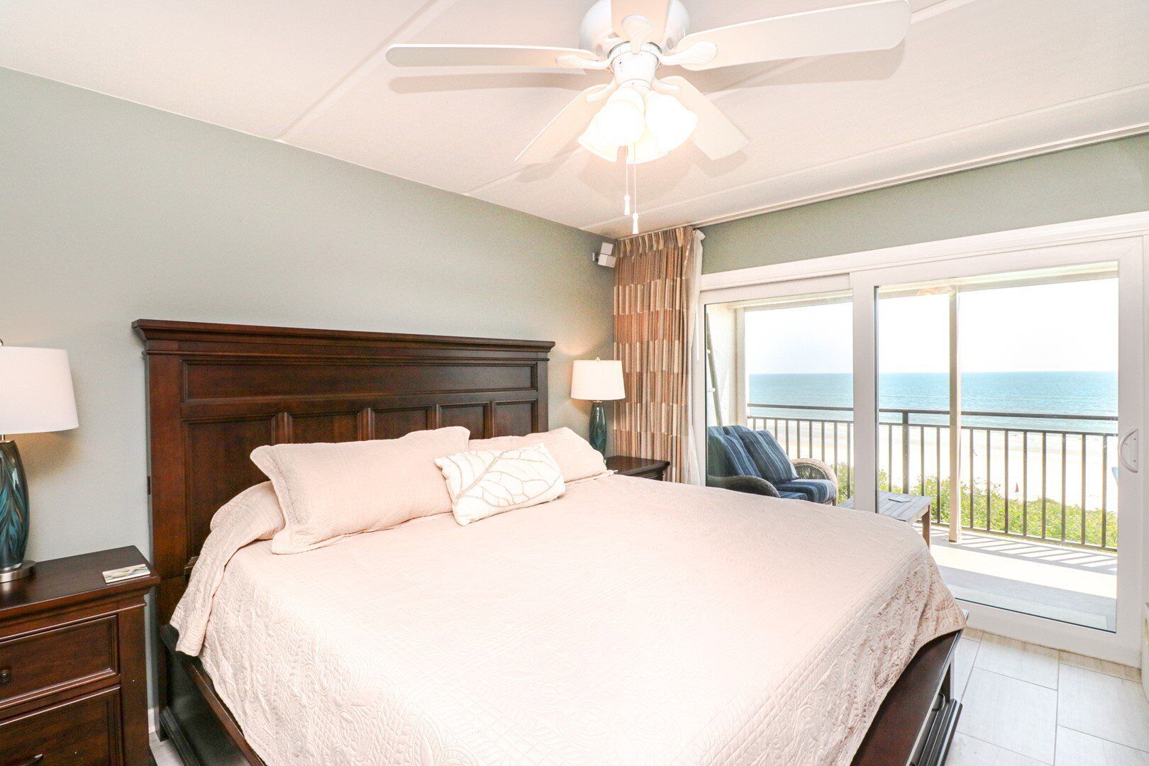 A bedroom with a large bed, a ceiling fan, a nightstand, a lamp, a chair, and a view of the ocean through a balcony.