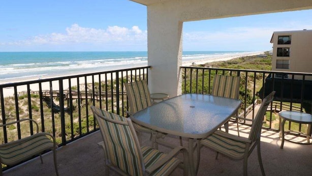 A balcony with a glass table and striped chairs, overlooking a beach and ocean. - Windjammer 312 - Remodeled, Direct Oceanfront Three Bedroom Crescent Beach Condo (St. Augustine)