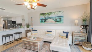 A modern living room with a white sofa, a coffee table, and a kitchen in the background. - Windjammer 404 - Updated, 4th Floor Oceanfront Condo with Elevator Access (St. Augustine)