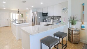A modern kitchen with white cabinetry, a central island with stools, stainless steel appliances, and a clock on the wall. - Windjammer 404 - Updated, 4th Floor Oceanfront Condo with Elevator Access (St. Augustine)