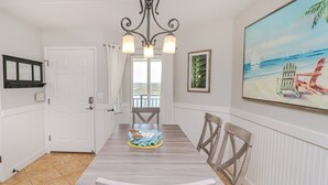 A dining area with a wooden table, chairs, a chandelier, and a painting of a beach scene. - Windjammer 404 - Updated, 4th Floor Oceanfront Condo with Elevator Access (St. Augustine)