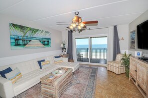 Een woonkamer met een witte bank, een salontafel, een televisie en uitzicht op het strand door een schuifdeur.