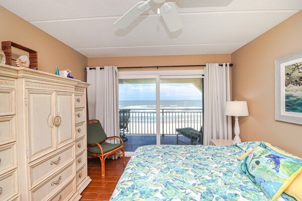 A bedroom with a bed, dresser, chair, and a balcony with a view of the ocean.