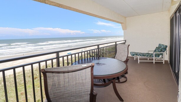 A balcony with a glass table and chairs, overlooking a beach with waves and a clear sky.