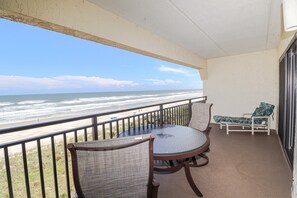 A balcony with a glass table and chairs, overlooking a beach with waves and a clear sky.