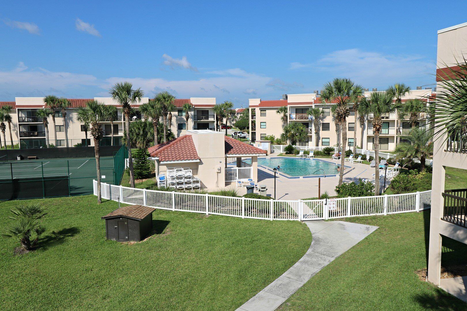 A resort with a swimming pool, tennis courts, and a fenced area with lounge chairs.