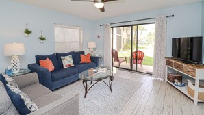 Ein Wohnzimmer mit blauem Sofa, einem Glastisch, einem Deckenventilator, einem Fernseher und Blick ins Freie.