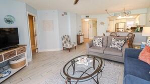 Ein Wohnzimmer mit Fernseher, Glastisch, Sofa und Sessel. Im Hintergrund ist eine Küche zu sehen.