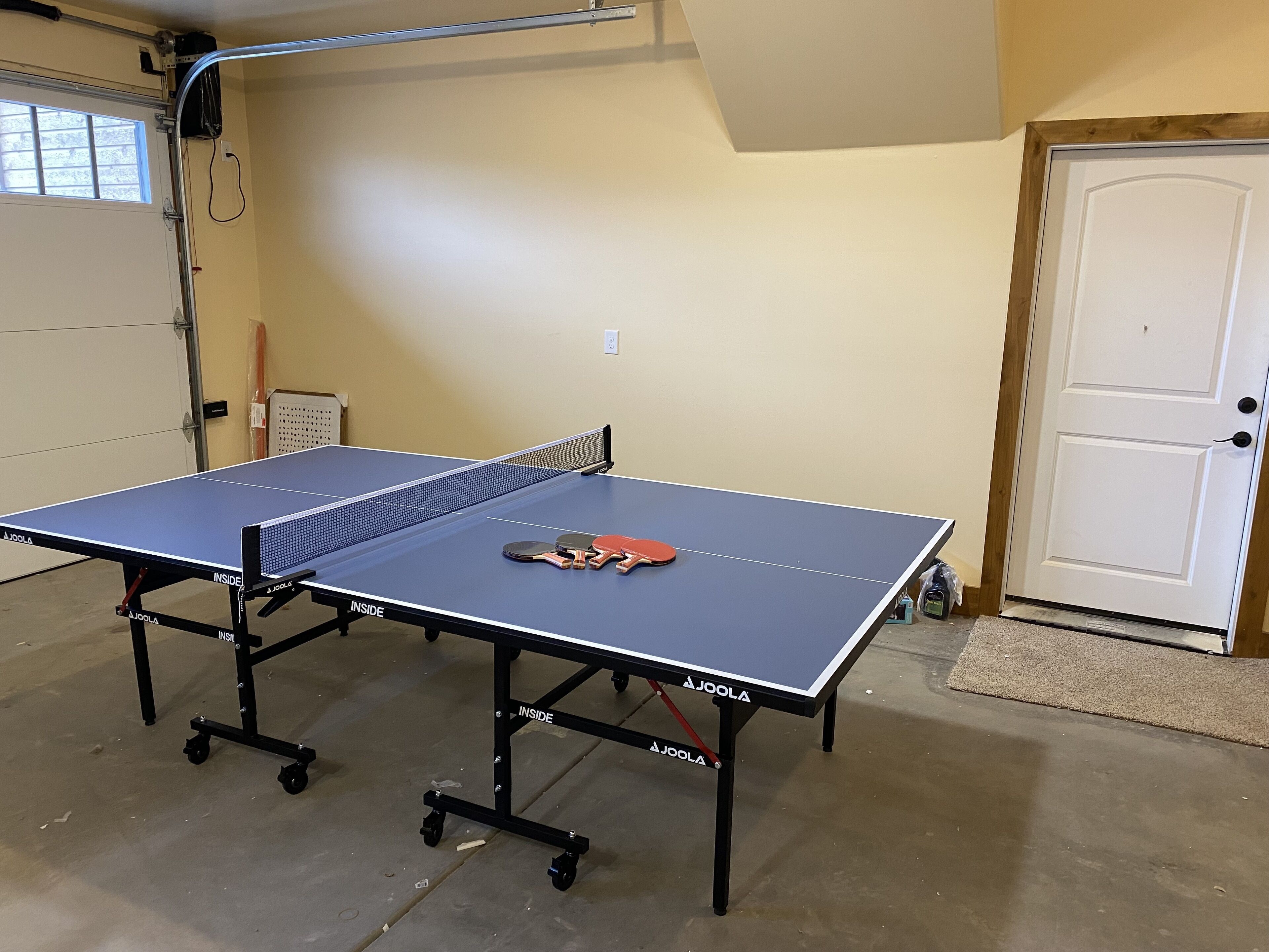 A ping pong table in a room with a door, a window, and a wall-mounted electrical outlet.
