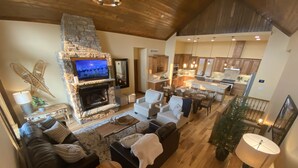 A spacious living area with a stone fireplace, a television, a dining area, and a kitchen visible through an open doorway. - Luxury Mountain Home in Grand Lake - Hot Tub (Grand Lake)