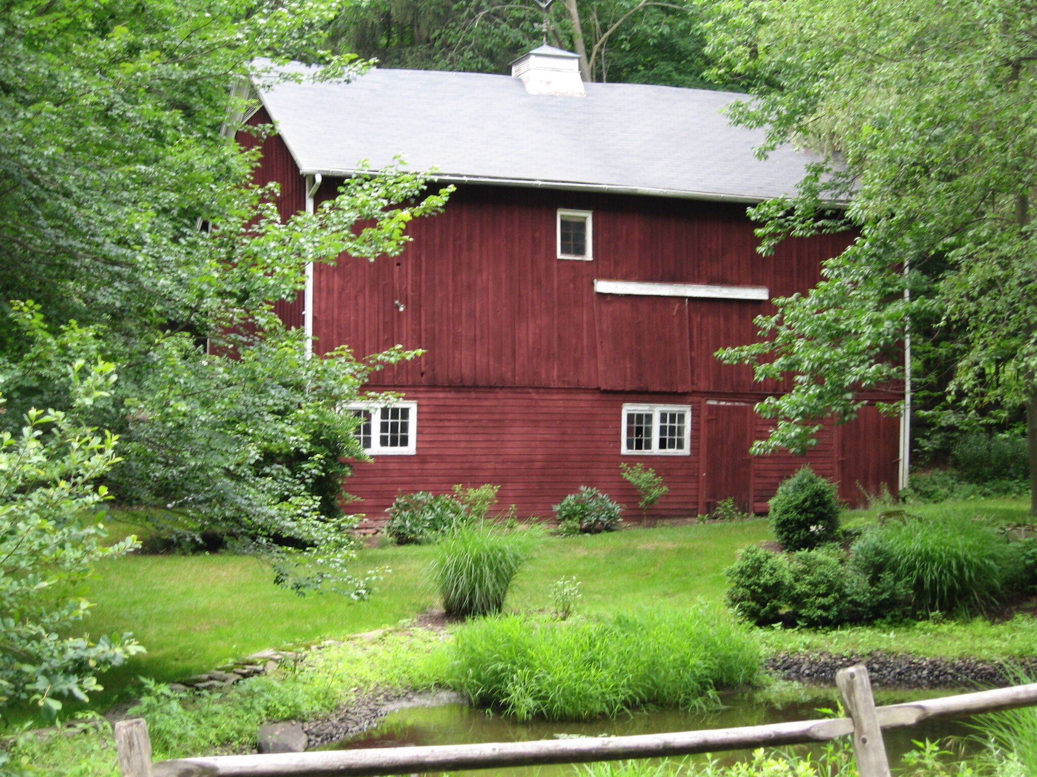 A red wooden house with a white window and door, surrounded by lush greenery and a small pond.