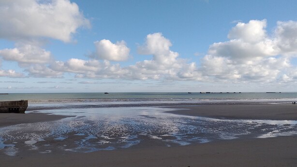 Ein Strand mit einer großen Betonkonstruktion, ruhigem Wasser und einem klaren Himmel mit Wolken.
