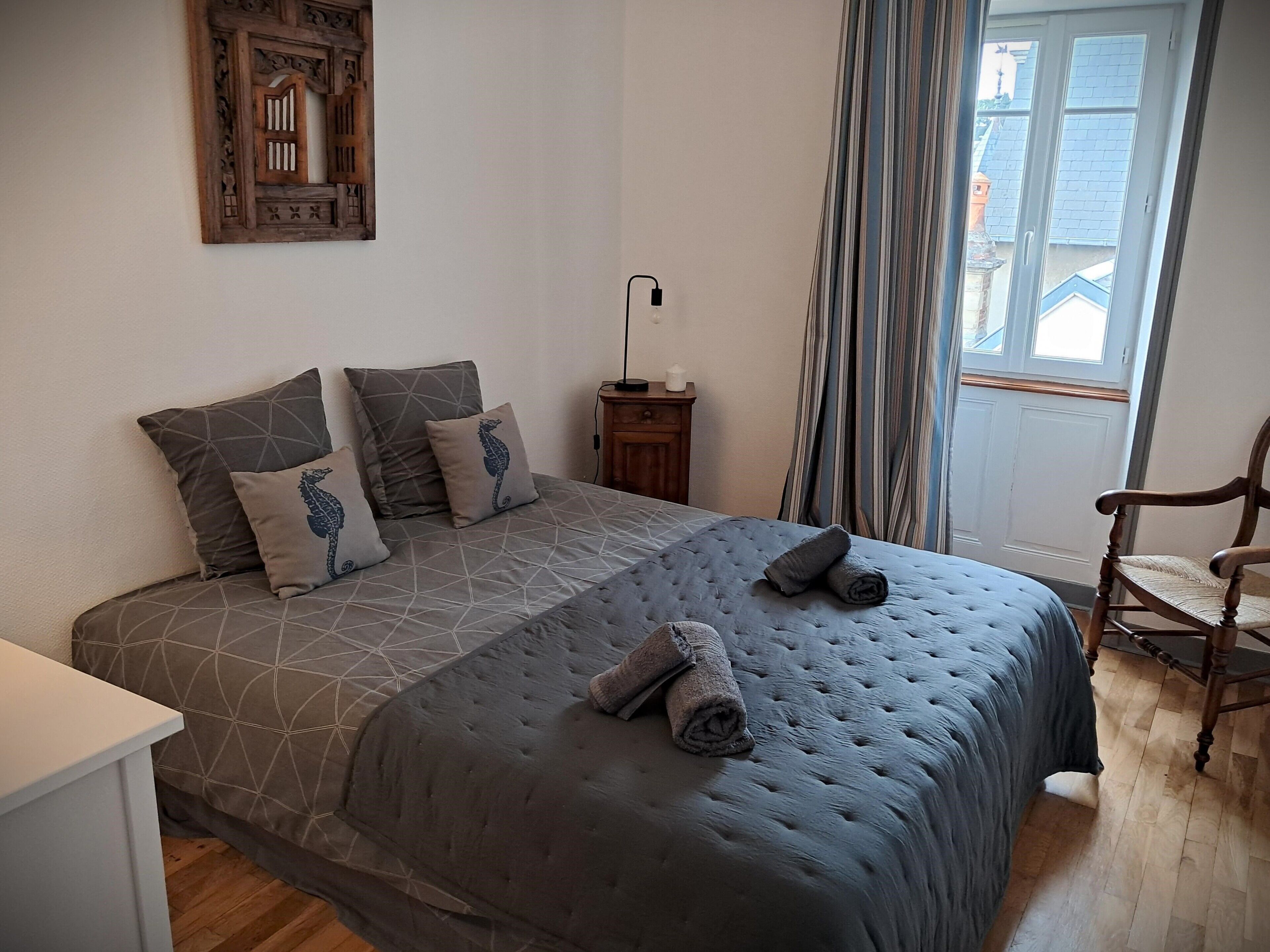 A bedroom with a large bed, a wooden chair, a nightstand, and a window with curtains.