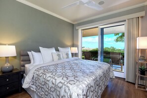 A bedroom with a large bed, two bedside tables with lamps, a ceiling fan, and a sliding glass door leading to an outdoor area with a chair and a plant. - Sleeps 10, Family Favorite, Fully Upgraded Modern Renovation and Decorated (Kihei)