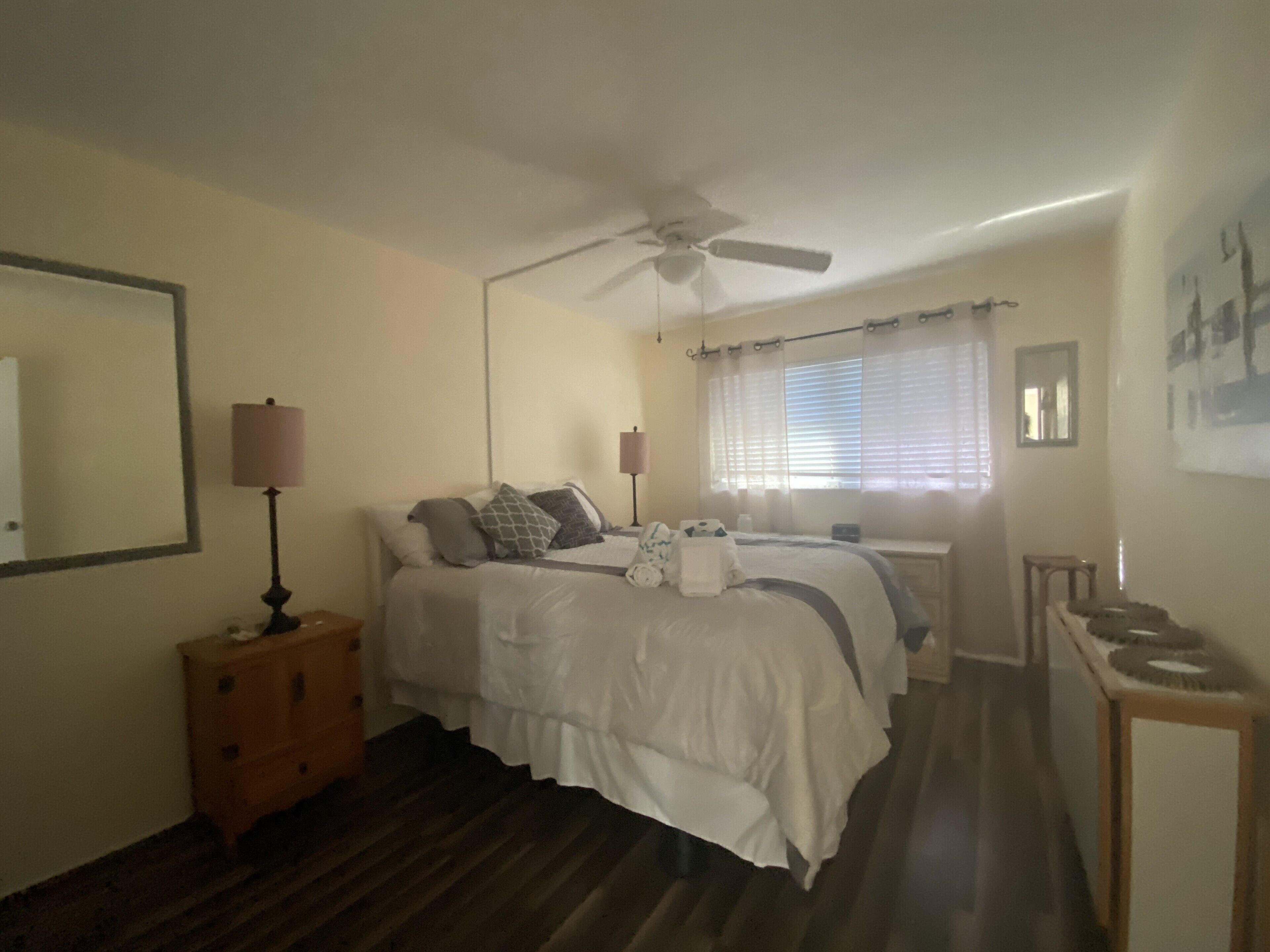 Quarto com cama, abajures, ventilador de teto, janela com persianas, mesa de cabeceira de madeira e espelho na parede.