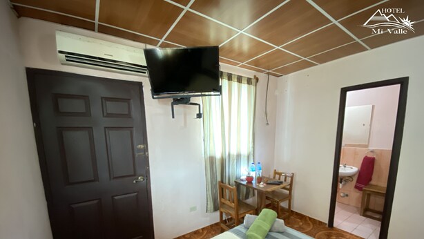 Habitación de hotel con cama, televisión montada en la pared, mesa de comedor con sillas y baño visible a través de una puerta abierta.
