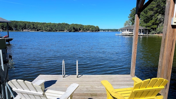 En träbrygga med två Adirondack-stolar, en vit och en gul, med utsikt över en stilla sjö och en skogbeklädd strand.