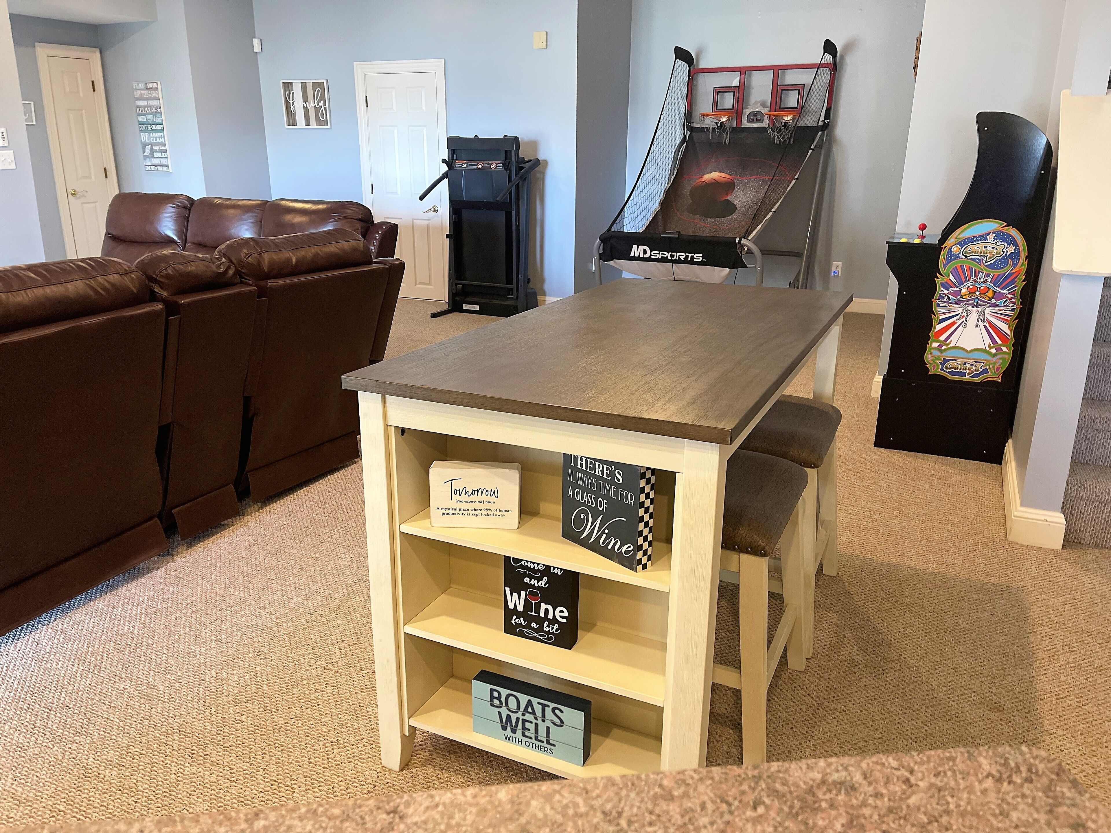 A living room with a brown leather couch, a wooden table with shelves, a treadmill, and a basketball hoop.