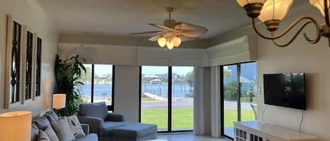 Una sala de estar con ventilador de techo, ventanas amplias, televisor y un mueble blanco.