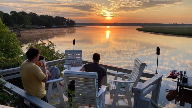 En træterrasse med hvide stole, der kigger ud over en rolig sø ved solnedgang.