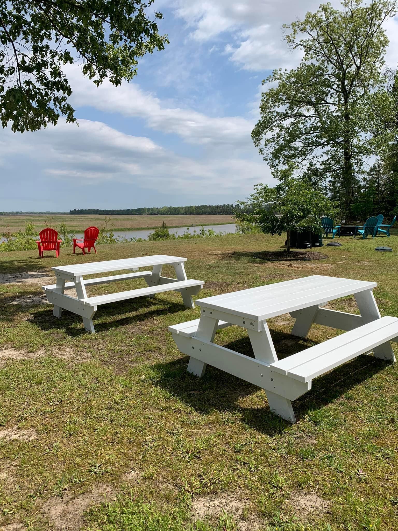 Due tavoli da picnic e diverse poltrone Adirondack in una zona erbosa, con un lago e alberi sullo sfondo.