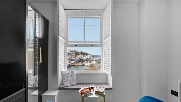 A modern room with a large window overlooking a harbor, a small round table with a book and a cup, and blue chairs.