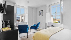 A modern hotel room with a bed, two blue chairs, a desk with a coffee machine, and a view of the outside.