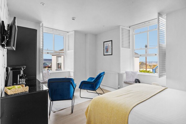 A modern hotel room with a bed, two blue chairs, a desk with a coffee machine, and a view of the outside.