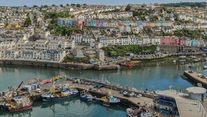 A coastal town with colorful buildings, a marina with boats, and a hilly backdrop.