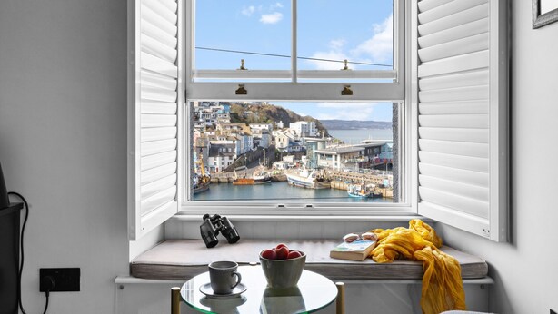A bay window with a view of a coastal town, a pair of binoculars, a bowl of fruit, and a book on a small table.