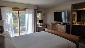 Ein Hotelzimmer mit einem großen Bett, einem Fernseher, einer hölzernen Bank, einem Schreibtisch mit Lampe und Blick auf Grünpflanzen durch das Fenster.
