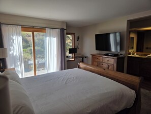 A hotel room with a large bed, a television, a wooden bench, a desk with a lamp, and a view of greenery through the window.