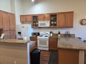 A kitchen with wooden cabinets, a microwave, a stove, a trash bin, and a clock on the wall.