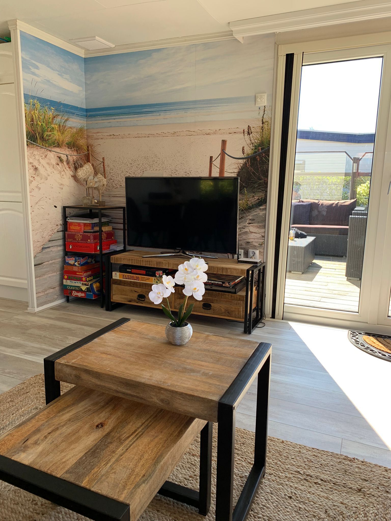 A living room with a beach mural, a flat-screen TV, a wooden coffee table, and a view of the outdoors.