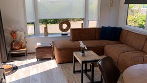 A modern living room with a brown sectional sofa, a wooden coffee table, a small side table, and a large window with a view of greenery.