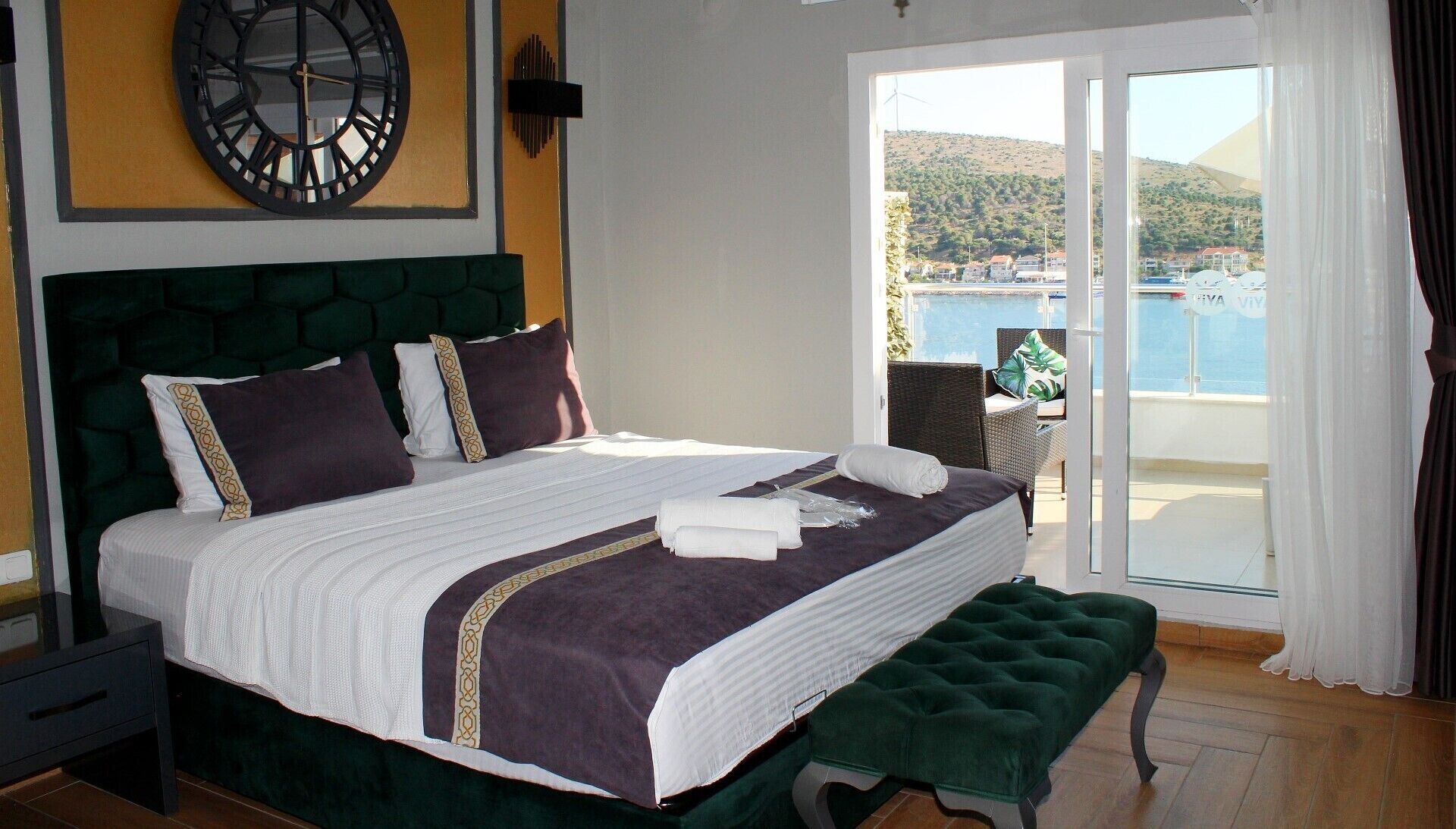 A hotel room with a large bed, a green headboard, a round wall clock, and a view of the sea through a sliding glass door.