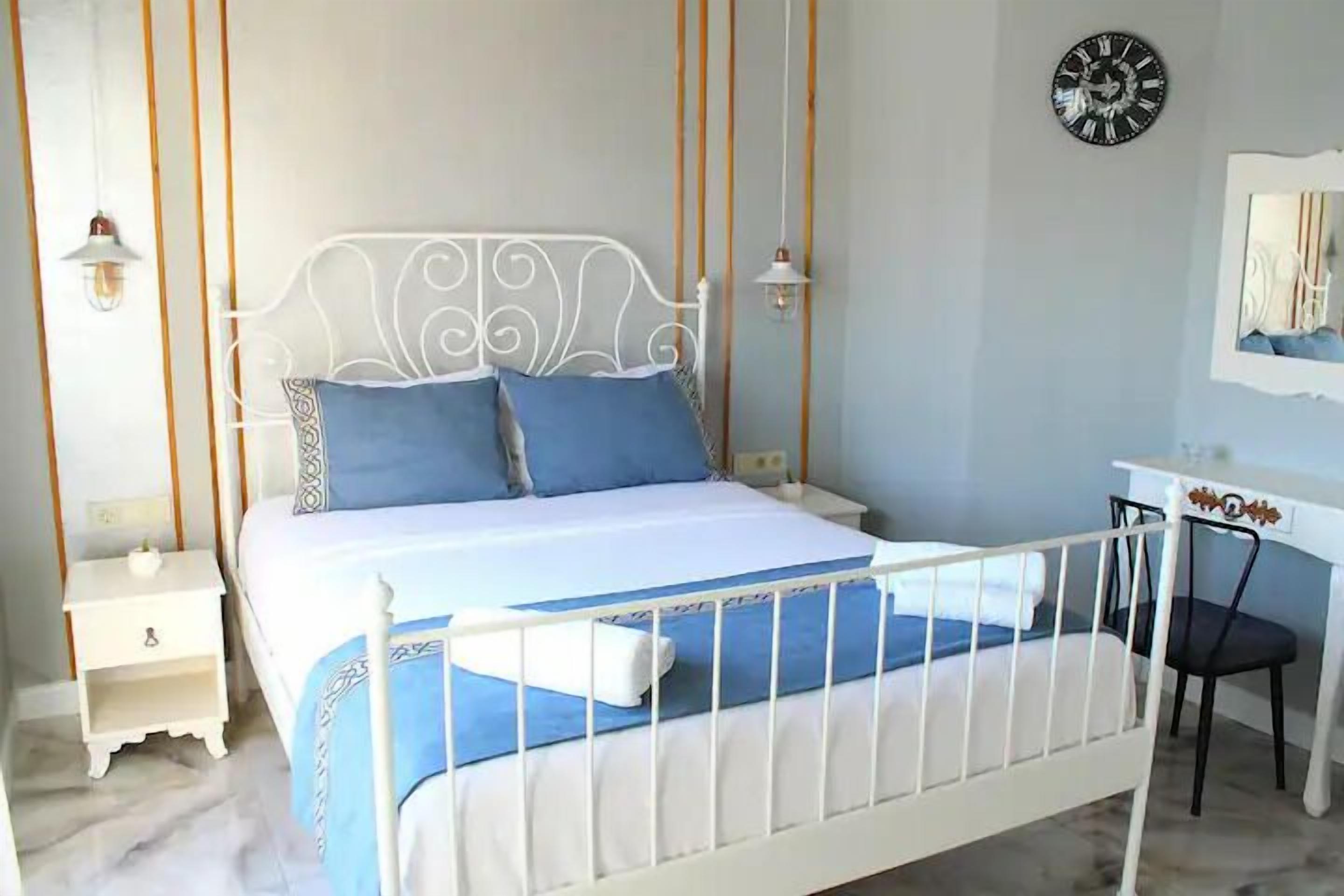 A bedroom with a white metal bed frame, a blue and white bedspread, two pillows, a bedside table with a lamp, and a wall clock.