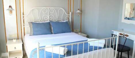 A bedroom with a white metal bed frame, a blue and white bedspread, two pillows, a bedside table with a lamp, and a wall clock.